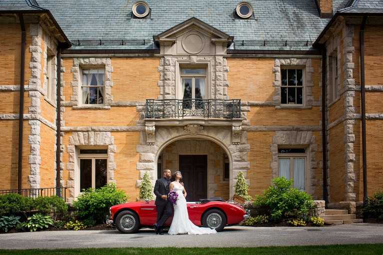 Bride and groom pose in the sun with red sports car in front of Cairnwood Estate in Bryn Athyn, PA